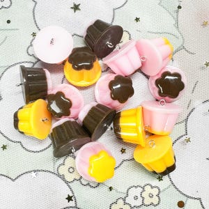 May include: A collection of cupcake-shaped charms in various colors, including pink, yellow, and brown. Each charm has a small loop for attaching to jewelry or other crafts. The charms are scattered on a patterned surface.