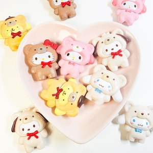 Può includere: Una collezione di figurine colorate in stile cartone animato, tra cui orsi, gatti e cani, sono disposte su un piatto rosa a forma di cuore. Ogni figurina è adornata con fiocchi e dettagliati tratti del viso, nelle tonalità del marrone, rosa, giallo e bianco.