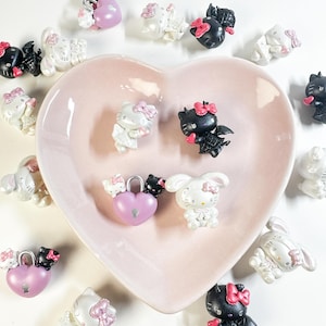 May include: A collection of small, decorative figurines featuring a popular cartoon character in various poses. The figurines are primarily white and black, with pink accents. Some are paired with heart-shaped lockets. The figurines are arranged around a pink heart-shaped dish.