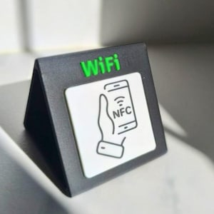 Puede incluir: Un letrero WiFi triangular negro con la palabra "WiFi" en verde. Un cuadrado blanco presenta una mano sosteniendo un teléfono con el símbolo NFC. El letrero está diseñado para la conectividad sin contacto.