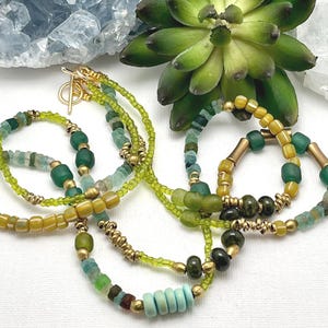 May include: A selection of beaded necklaces and bracelets in green, yellow, and blue hues. The jewellery features diverse bead shapes and sizes, with gold-coloured metal accents. A blue crystal geode and a green succulent are in the background.