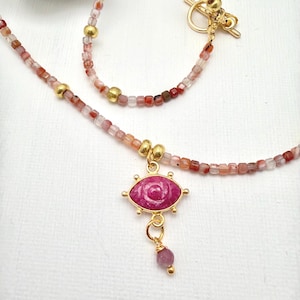 May include: A delicate necklace featuring a pink gemstone eye pendant with gold accents. The necklace is made of small, square, pink and red beads interspersed with gold beads. The necklace has a gold toggle clasp.