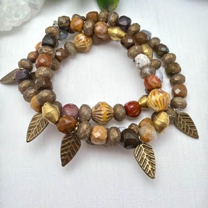 Puede incluir: Tres pulseras de cuentas con cuentas en tonos tierra, marrón, dorado y rojo. Cada pulsera tiene colgantes de hojas doradas. Las pulseras están dispuestas sobre una superficie blanca. Las cuentas se miden en centímetros.
