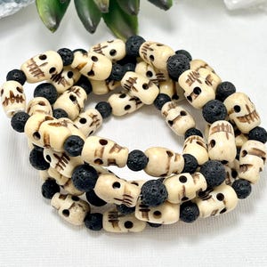 May include: A necklace featuring off-white skull beads interspersed with black lava rock beads. The skull beads have carved facial details. The necklace is displayed against a backdrop of a green succulent plant and a white, crystalline object.