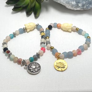 Puede incluir: Dos pulseras de cuentas con una variedad de piedras coloridas. Una pulsera tiene un dije de loto plateado, la otra un dije de loto dorado. Ambas pulseras presentan una cabeza de Buda color crema y detalles plateados. Las pulseras se exhiben sobre una superficie blanca.
