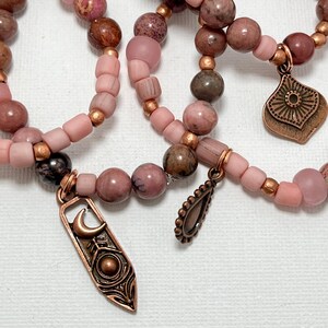 Puede incluir: Primer plano de tres pulseras de cuentas en tonos rosa, marrón y cobre. Las pulseras presentan varias formas y tamaños de cuentas, con espaciadores de color cobre y dijes decorativos. Los dijes incluyen una media luna y un diseño de lágrima.