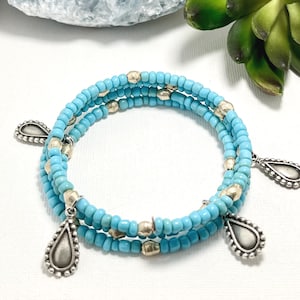 Puede incluir: Una pulsera envolvente con tres hilos de cuentas azul claro y detalles plateados. La pulsera presenta colgantes plateados en forma de lágrima y cuentas plateadas. La pulsera se exhibe sobre una superficie blanca.