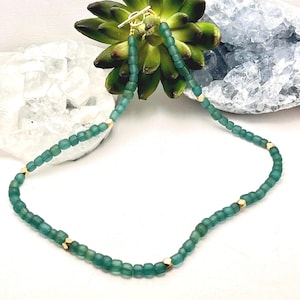Op de afbeelding: Een turquoise kralenketting met goudkleurige accenten. De ketting heeft kleine, vierkante kralen en goudkleurige, geometrisch gevormde kralen. De ketting wordt gepresenteerd op een wit oppervlak met een groene vetplant en een blauw kristal op de achtergrond.