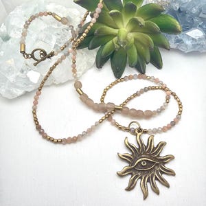 May include: A necklace featuring a bronze sun pendant with a central eye design. The chain consists of small, faceted peach-coloured beads interspersed with metallic gold-toned spacer beads and a bronze toggle clasp closure at the back.