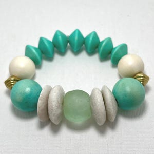 May include: A turquoise and cream-coloured beaded bracelet. The bracelet features turquoise diamond-shaped beads, cream-coloured round beads, and light green and white stone beads. Gold-coloured metal accents are also present.