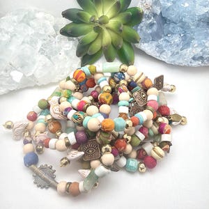 Op de afbeelding: Een verzameling kralenarmbanden in diverse kleuren, waaronder turquoise, oranje en roze. De armbanden hebben houten kralen, stoffen accenten en metalen bedels met oogontwerpen. Sommige armbanden hebben goudkleurige metalen accenten.