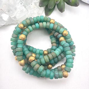 May include: A necklace composed of turquoise-colored, cylindrical beads interspersed with gold-colored spherical beads. The beads are arranged in a spiral pattern, creating a layered effect. The necklace is displayed on a white surface.