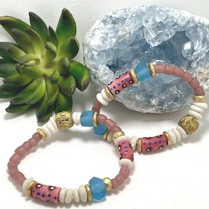 Peut inclure: Deux bracelets de perles. L'un présente des perles roses, blanches et dorées, avec des perles bleues. L'autre bracelet a des perles similaires. Les bracelets sont exposés sur une surface blanche.
