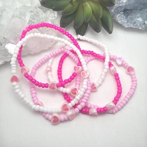 Op de afbeelding: Een verzameling kralenarmbanden in roze en witte tinten. Elke armband heeft kleine hartvormige accenten. De armbanden zijn in een gelaagde opstelling gerangschikt, die de verscheidenheid aan kleuren en ontwerpen laat zien.