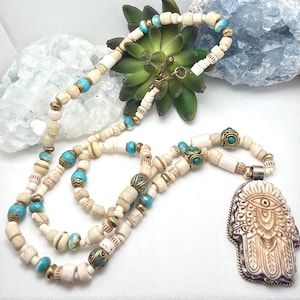 May include: A cream-colored beaded necklace with turquoise and gold accents. The necklace features a toggle clasp and a decorative Hamsa hand pendant. The beads are a mix of cylindrical and round shapes, creating a bohemian style.