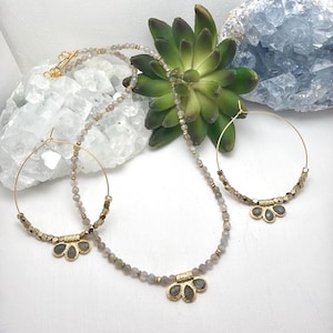 May include: A jewelry set featuring a gold necklace, earrings, and a gold clasp. The necklace and earrings have small, round, gray beads and gold accents. The earrings are hoop style. The set is displayed with a green succulent and crystals.