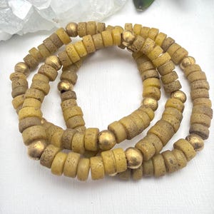 Può includere: Una collana con perline cilindriche verde oliva e perline sferiche dorate. Le perline sono infilate insieme, creando un effetto a strati. La collana è esposta su uno sfondo bianco.