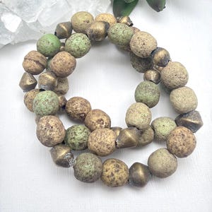 May include: A necklace featuring a strand of textured, round beads in shades of green, tan, and brown, interspersed with metallic cone-shaped beads. The beads have a speckled appearance, creating a rustic aesthetic.