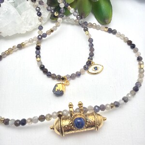 May include: A collection of beaded necklaces with gold accents and various pendants. The necklaces feature small, round beads in shades of gray, purple, and gold. Pendants include an eye charm, a stone with gold leaf, and a gold cylinder with a blue stone.