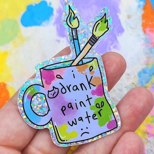 May include: A holographic sticker featuring a cartoon mug with paintbrushes. The mug is filled with colorful paint and the words "i drank paint water" are written on it. The sticker has a sad face at the bottom.