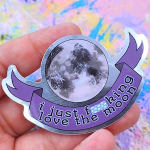 May include: A silver sticker featuring a full moon graphic with a purple banner. The banner reads "i just f*cking love the moon" in white text. The sticker is held in a hand.