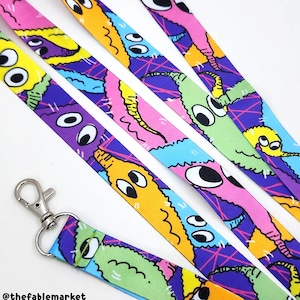 A neon colored lanyard featuring a vivid print of fuzzy pipe cleaner inspired worm creatures. The worms are all entangled with one another and have big, mismatched eyes giving them a charming silly look. The lanyard has a silver hook.