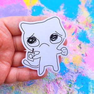 May include: A white sticker featuring a sad cartoon cat holding a drink and a cookie. The cat has large, scribbled eyes and a downturned mouth. The sticker is held against a colorful, abstract background.