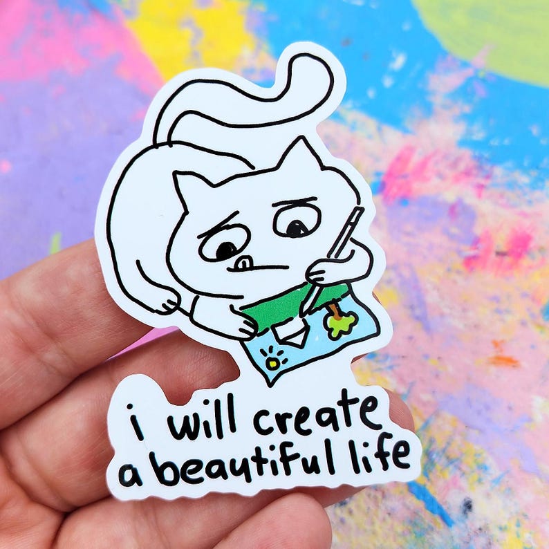 May include: A white sticker featuring a cartoon cat drawing on a green and blue paper. The cat has a sad expression and is holding a pencil. The sticker has the text "I will create a beautiful life."