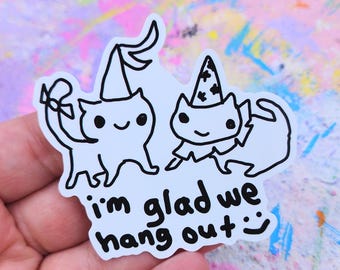 May include: A white sticker featuring two cartoon cats with party hats. The cats are outlined in black and have smiling faces. The text "I'm glad we hang out :)" is written below the cats in black.