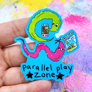 May include: A blue sticker featuring a cartoon worm with a green head and pink body. The worm is holding a phone and a drawing. The sticker reads "parallel play Zone" with star accents.