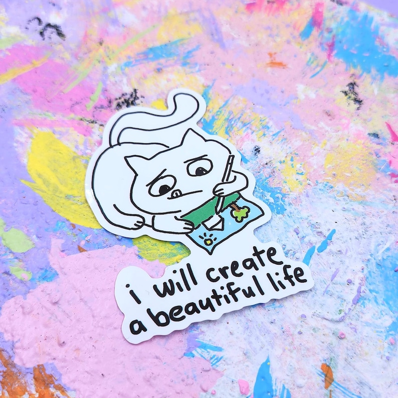 May include: A white cat sticker with a black outline, holding a pen and drawing on a blue paper. The sticker has the text "i will create a beautiful life" in black. The background is a colorful mix of pink, purple, and blue paint.