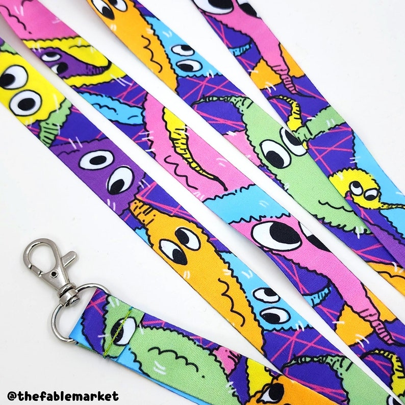 A neon colored lanyard featuring a vivid print of fuzzy pipe cleaner inspired worm creatures. The worms are all entangled with one another and have big, mismatched eyes giving them a charming silly look. The lanyard has a silver hook.