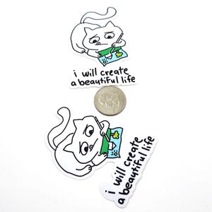 May include: White cat stickers with black outlines, each depicting a cat drawing a picture. The picture includes a house, tree, and water. The text "i will create a beautiful life" is below the cat. A quarter is next to the stickers.
