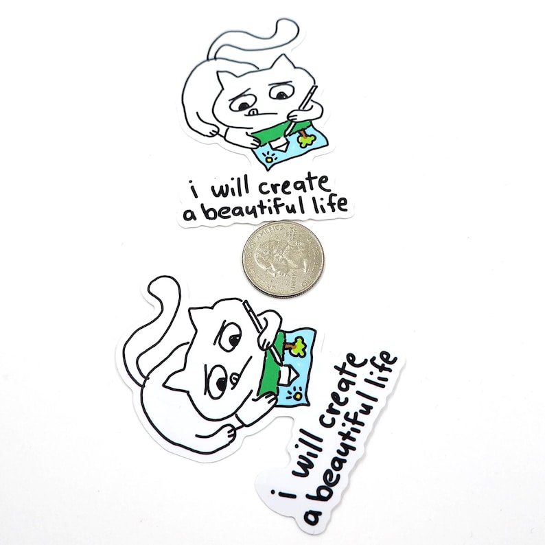 May include: White cat stickers with black outlines, each depicting a cat drawing a picture. The picture includes a house, tree, and water. The text "i will create a beautiful life" is below the cat. A quarter is next to the stickers.