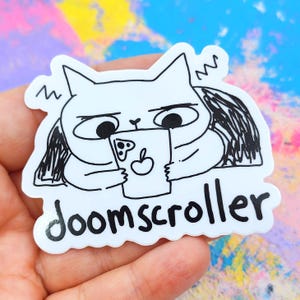 May include: A white sticker featuring a cartoon cat holding a phone with an apple logo. The cat has a grumpy expression and the word "doomscroller" is written below in black text. The sticker is held in a hand against a colorful background.