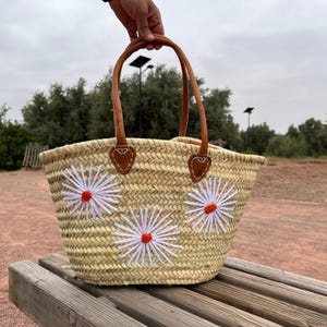オレンジと白の花があしらわれた、パーソナライズされたモロッコ風ストローバスケット。レザーハンドルと巾着付きのハンドメイドトートバッグ。