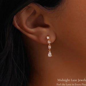 May include: A gold-toned earring with three clear, sparkling stones. The top two stones are square, and the bottom stone is a teardrop shape. The earring is dangling from an ear. Text at the bottom reads "Midnight Luxe Jewelry" and "Feel the Love in Every Piece."