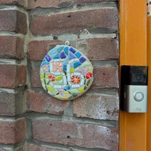 Peut inclure: Une plaque de mosaïque ronde avec le numéro "55" au centre, fixée à un mur de briques. La mosaïque présente des carreaux de verre colorés bleus, roses, verts et rouges. Une sonnette Ring est montée sur le cadre de porte adjacent.