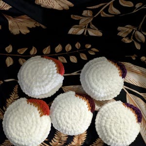 May include: Six white, round, crocheted items with a colorful accent on one side. The accent colors include shades of orange, brown, and purple. The items are arranged on a black background with a floral pattern.