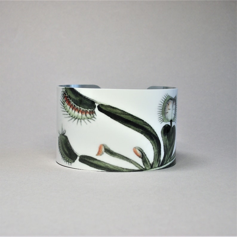Pu&ograve; includere: Bracciale a polsino bianco con un'illustrazione botanica di una Venere acchiappamosche. Il design include foglie verdi, trappole con punte rosse e accenti arancioni. L'interno del bracciale &egrave; color argento.