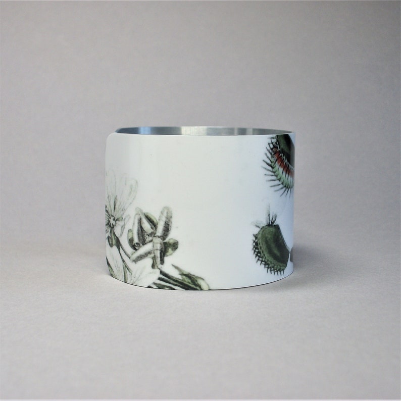 Pu&ograve; includere: Un braccialetto cilindrico bianco con interno argentato. Il braccialetto &egrave; decorato con illustrazioni botaniche di piante, tra cui una dionea e altri fiori in tonalit&agrave; di verde e nero.