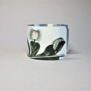Pu&ograve; includere: Bracciale a polsino in ceramica bianca con un'illustrazione botanica di una Venere acchiappamosche. Il design presenta le trappole e le foglie della pianta in verde e nero, con un bordo in metallo argentato.