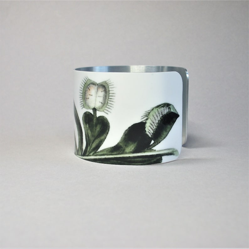 Pu&ograve; includere: Bracciale a polsino in ceramica bianca con un'illustrazione botanica di una Venere acchiappamosche. Il design presenta le trappole e le foglie della pianta in verde e nero, con un bordo in metallo argentato.