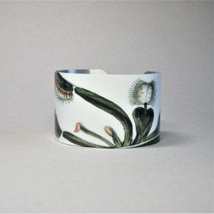 Pu&ograve; includere: Bracciale a polsino bianco con illustrazione botanica di una Venere acchiappamosche. Il design presenta foglie verdi, trappole con punte rosse e una fascia in metallo argentato. Un accessorio unico.