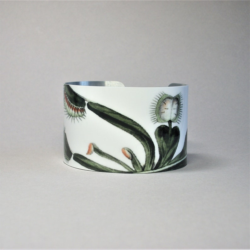Pu&ograve; includere: Bracciale a polsino bianco con illustrazione botanica di una Venere acchiappamosche. Il design presenta foglie verdi, trappole con punte rosse e una fascia in metallo argentato. Un accessorio unico.