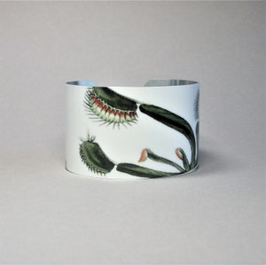 Pu&ograve; includere: Bracciale a polsino bianco con un'illustrazione botanica di una Venere acchiappamosche. Il design presenta foglie verdi, trappole rosse e bianche e una fascia in metallo argentato. Un accessorio unico.