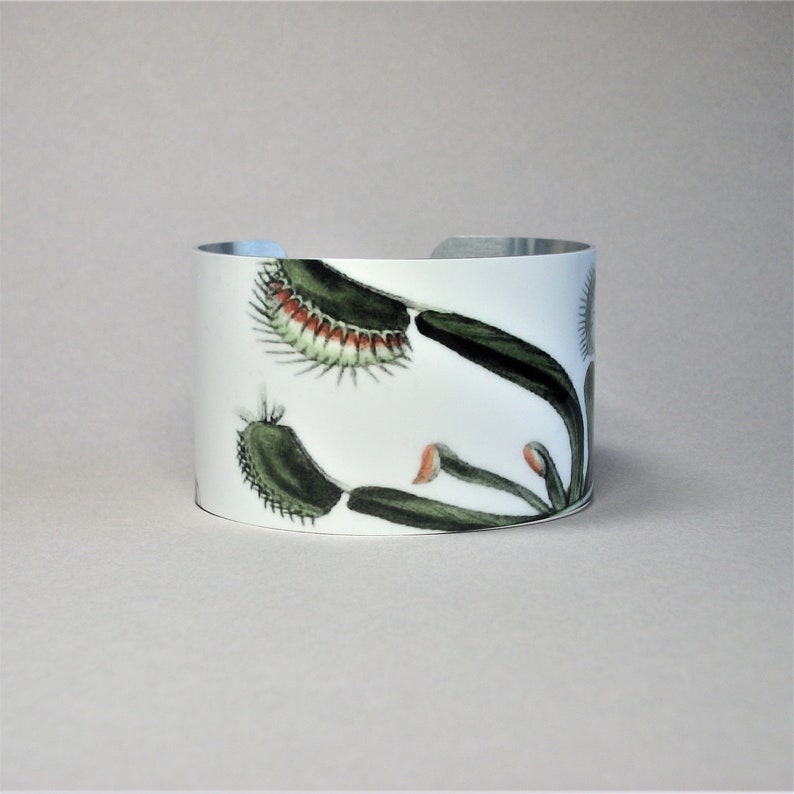 Pu&ograve; includere: Bracciale a polsino bianco con un'illustrazione botanica di una Venere acchiappamosche. Il design presenta foglie verdi, trappole rosse e bianche e una fascia in metallo argentato. Un accessorio unico.
