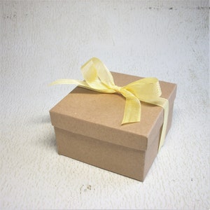 Pu&ograve; includere: Una piccola scatola regalo quadrata in cartone marrone, legata con un nastro giallo pallido. La scatola &egrave; chiusa e il nastro &egrave; legato con un fiocco sulla parte superiore. Lo sfondo &egrave; una superficie strutturata bianco sporco.