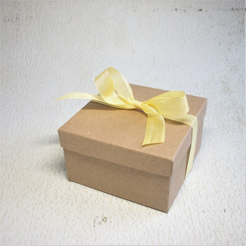 Pu&ograve; includere: Una piccola scatola regalo quadrata in cartone marrone, legata con un nastro giallo pallido. La scatola &egrave; chiusa e il nastro &egrave; legato con un fiocco sulla parte superiore. Lo sfondo &egrave; una superficie strutturata bianco sporco.