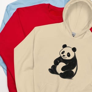 May include: A cream-colored hoodie featuring a black and white panda bear illustration. The hoodie has a kangaroo pocket and a drawstring hood. A red and a light blue crewneck sweatshirt are visible in the background.